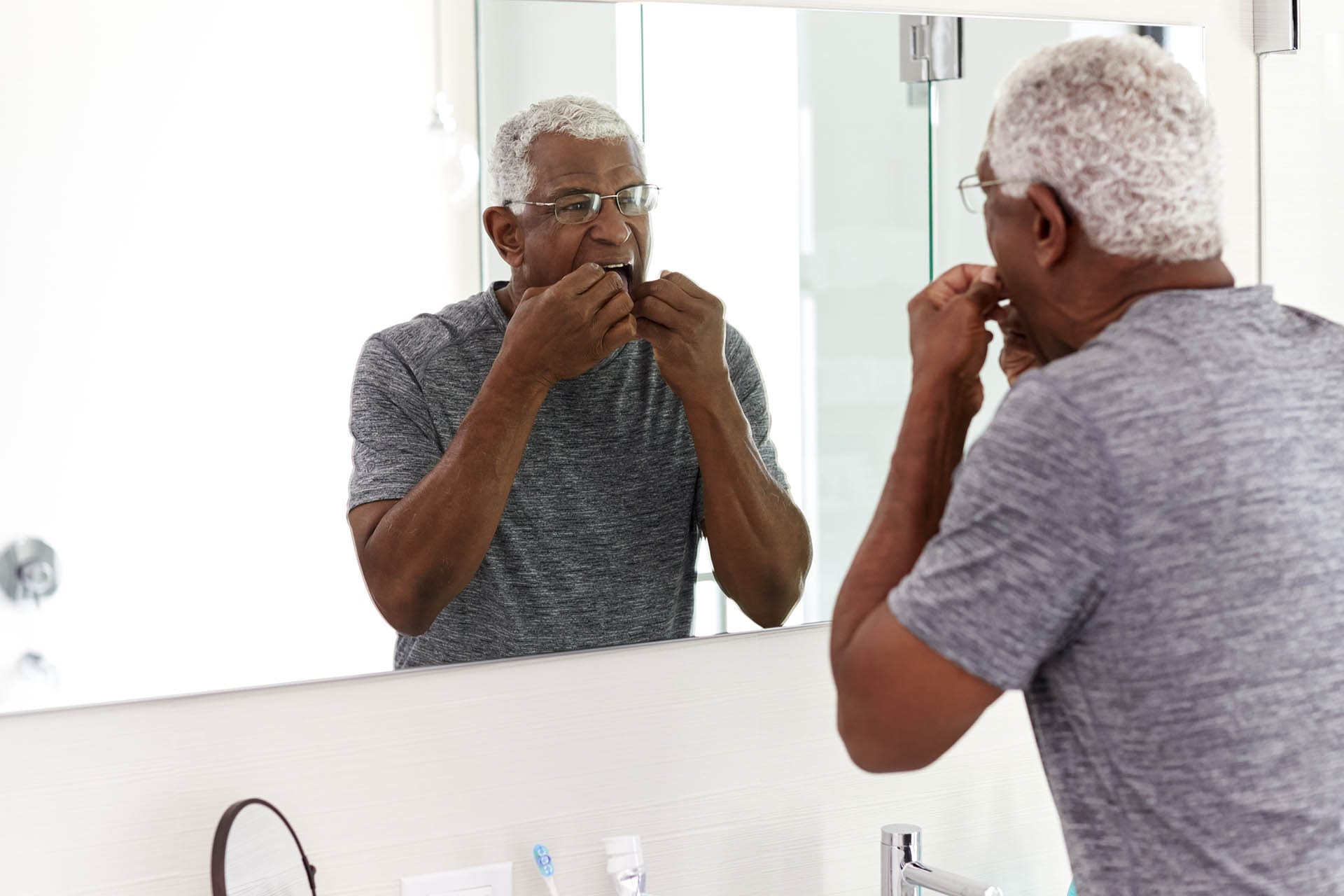 Senior Man Flossing Teeth Looking At Reflection In Bathroom Mirror Wearing Pajamas
